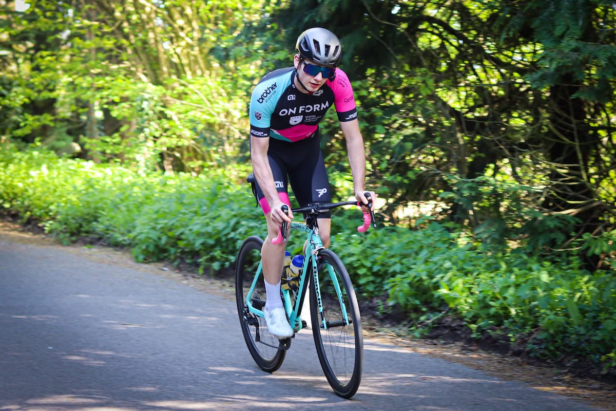 Cyclist Corey Bale standing and leaning forward while riding along a tree lined country road
