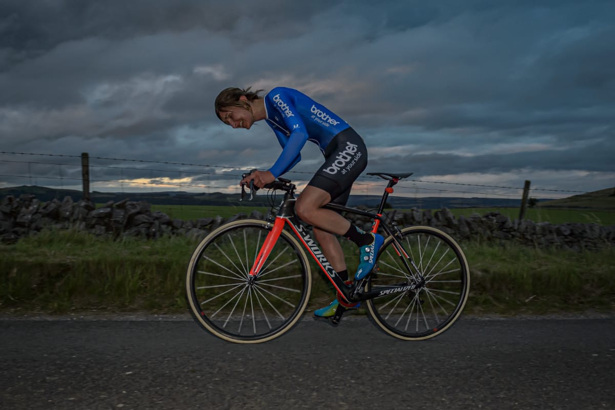 Side shot of cyclist Rebecca Richardson leaning over her handlebars as she rides along a country road with greenery and grey sky in the background