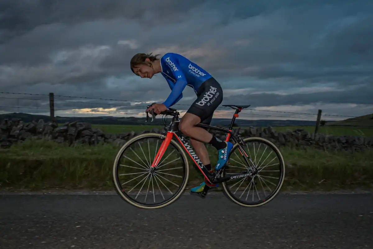 Side shot of cyclist Rebecca Richardson leaning over her handlebars as she rides along a country road with greenery and grey sky in the background