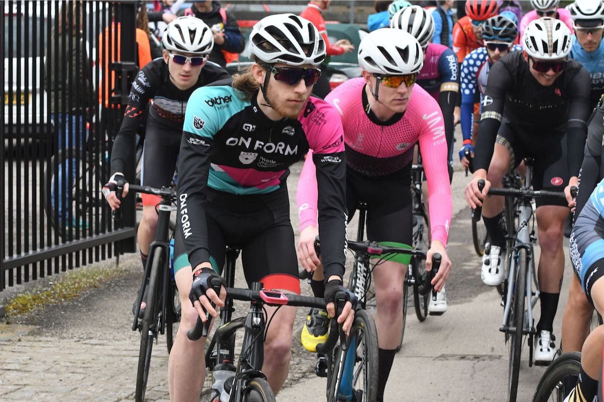 A group of cyclists leaning into a bend after riding past a black metal gate with spectators in the background