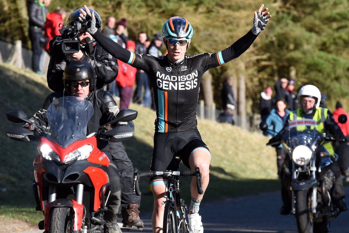 Cyclist Erick Rowsell riding with his arms in the air while being flanked by motorcyclists with spectators in the background