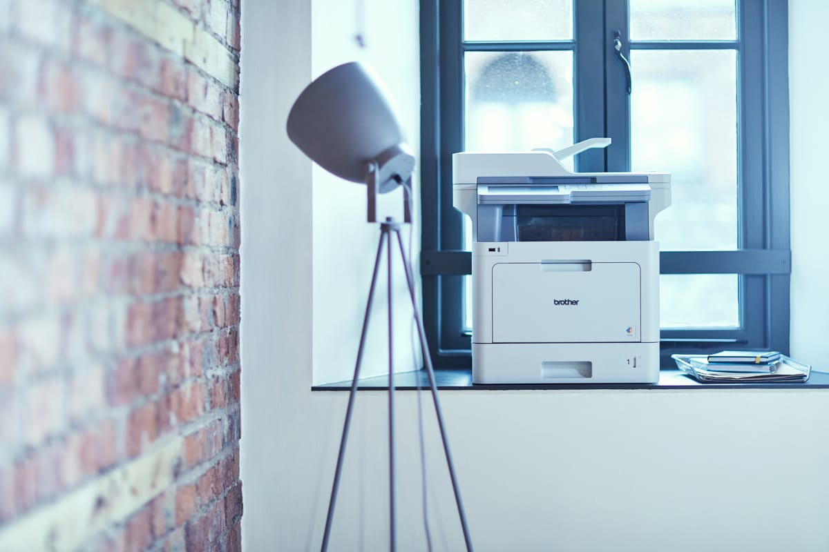 Brother MFC colour laser printer on a window ledge next to a tripod floor lamp