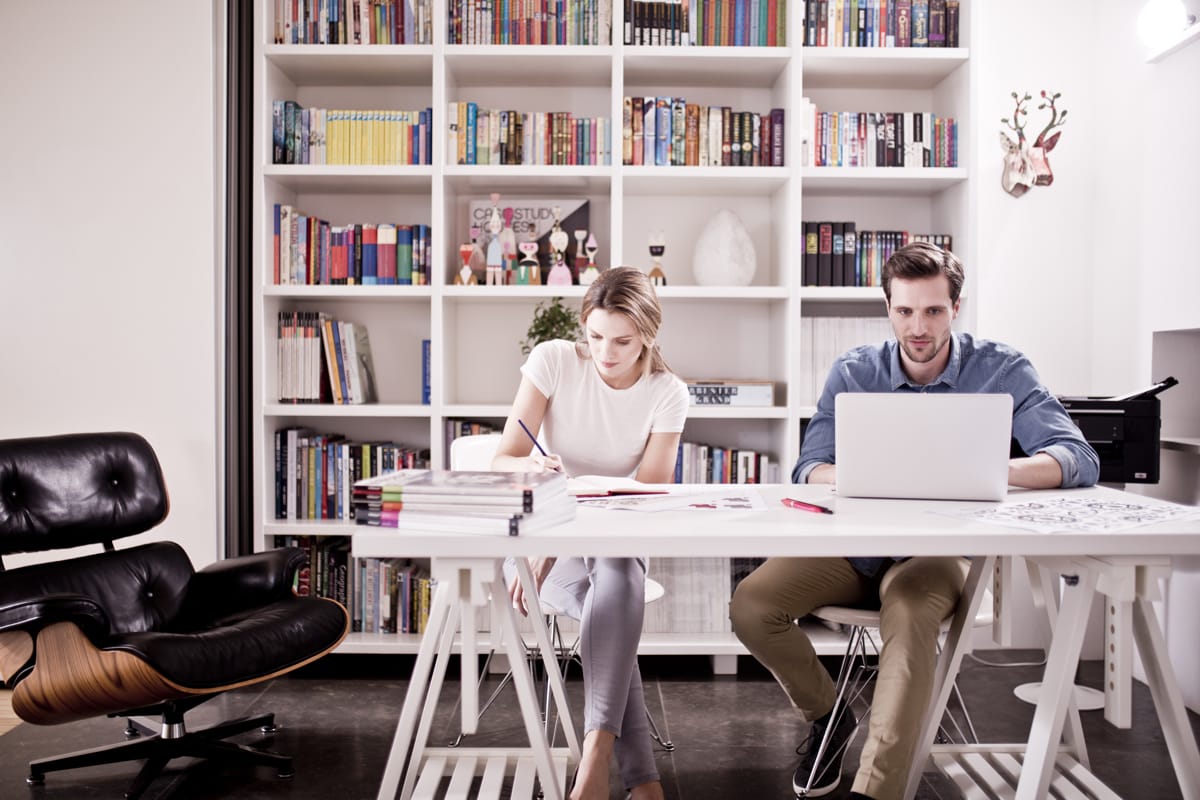 A lady and gentleman working at a desk in a home office with a bookcase in the background