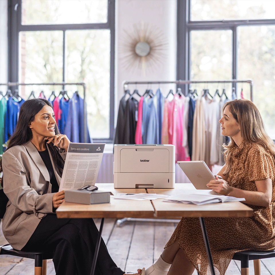 Two women in retail store sat at table with Brother HL-L5215DN, clothes rail, clothes