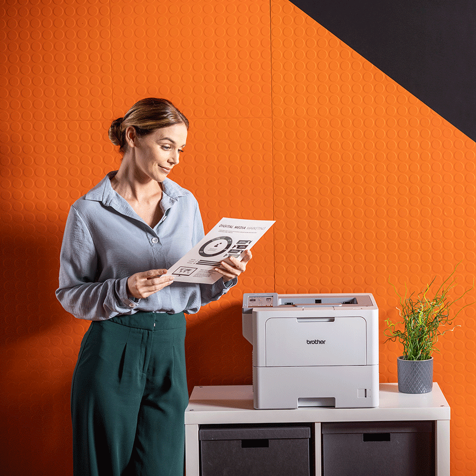 Female stood holding mono document next to Brother HL-L6415DN printer, plant, orange wall