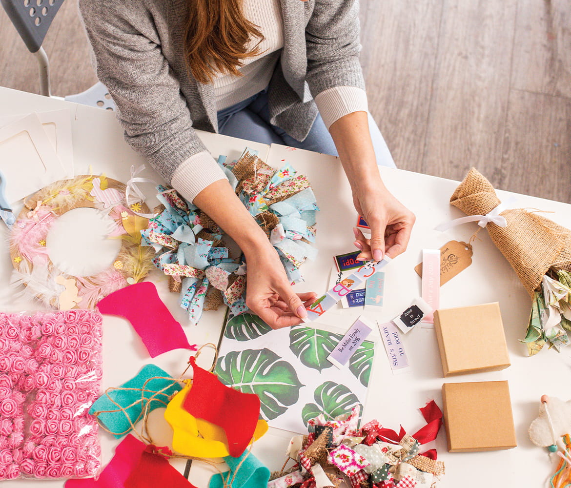 Woman sat at a table holding a label. The table is covered with paper designs including wreaths and flowers.