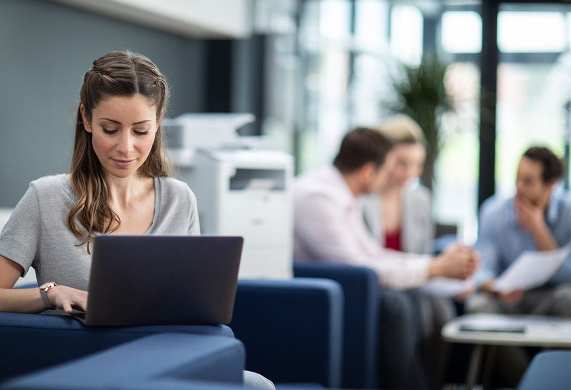 Woman sat on sofa using laptop with people having meeting in background