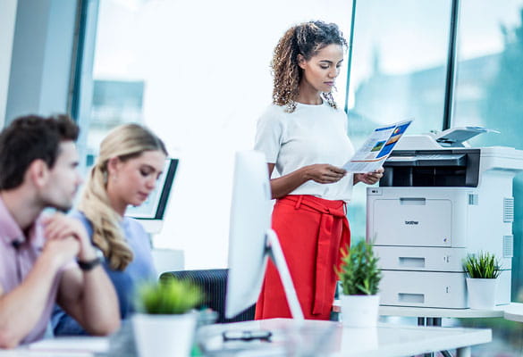 Man and woman sat at desk, female stood at printer, colour documents, table, plants