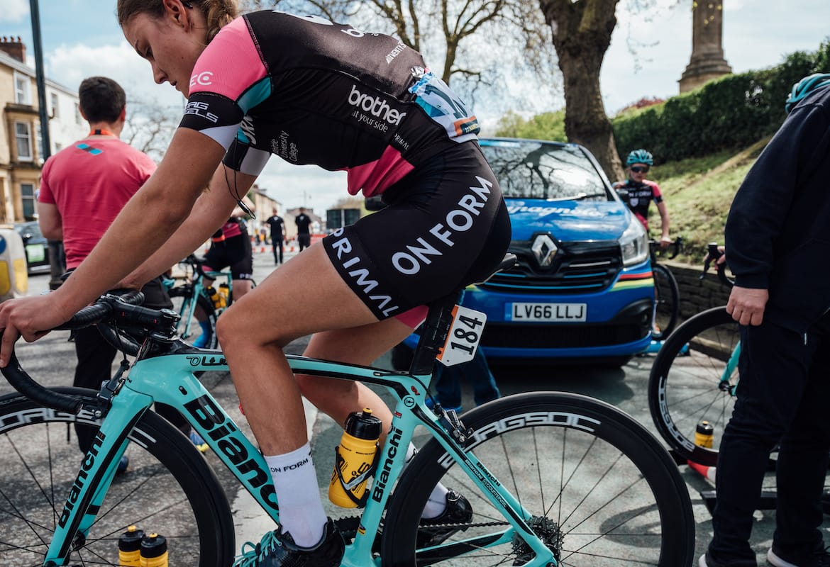 Side view of Team OnForm cyclist on a cordoned off road with other team members and a blue car in the background