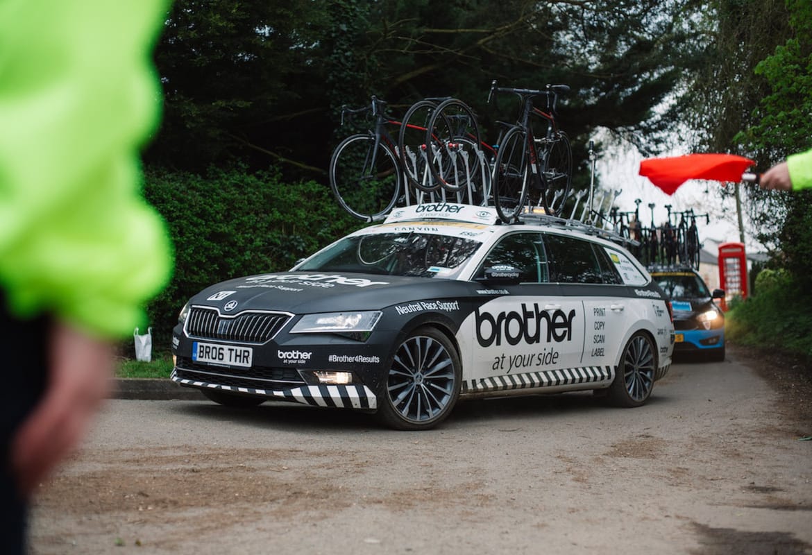 Brother cycling neutral support car turning into a bend with race marshals partially visible in the foreground and another support vehicle following in the background