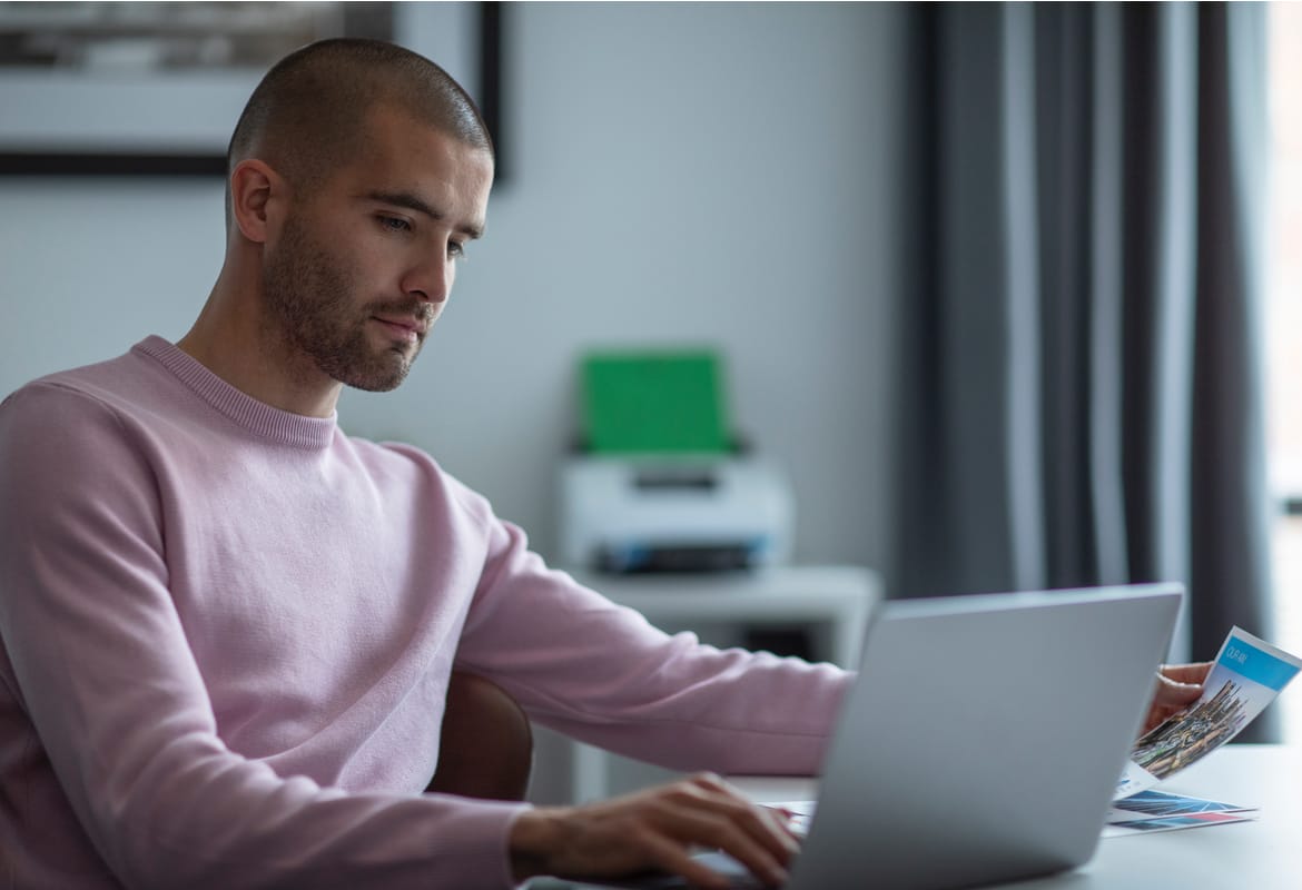 A man wearing a pink jumper using a notebook computer while sat at desk with a Brother ADS-4700W desktop document scanner in the background