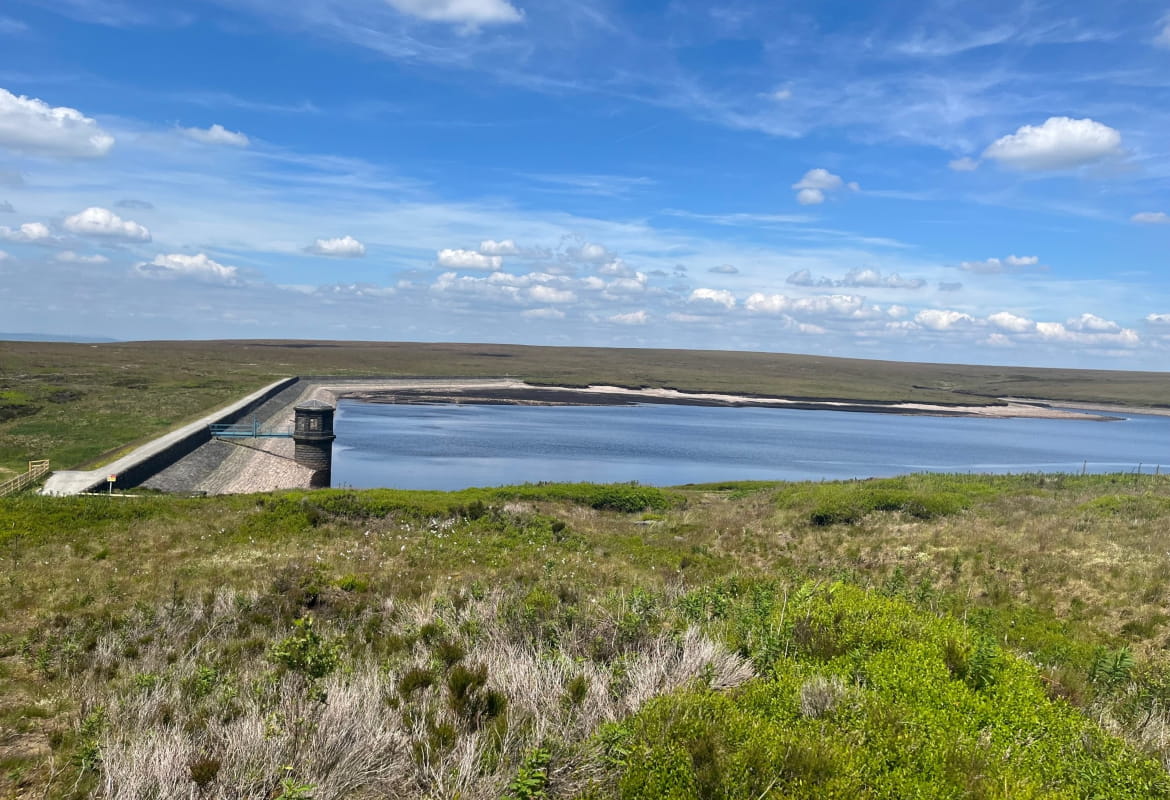 Dovestone reservoir filled with water surrounded by green grass and a blue sky with clouds