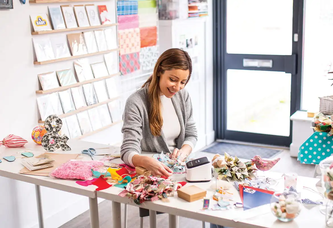 Femmes assises à une table dans une devanture de magasin imprimant des étiquettes, entourées de ciseaux, de cartes et de boîtes.