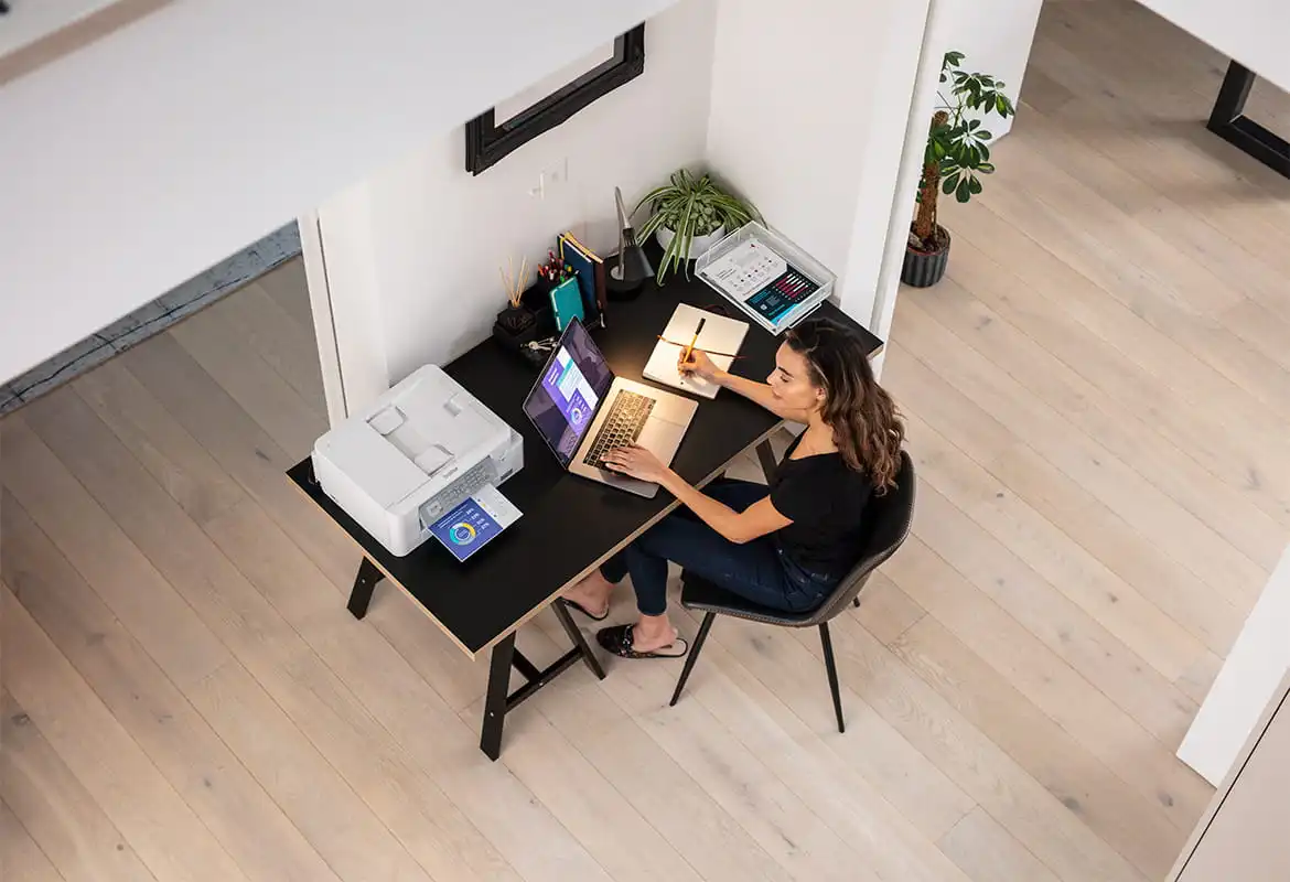 Femme assise à un bureau, regardant un ordinateur portable et une imprimante à côté.