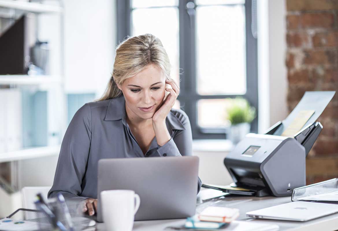 Woman with long blond hair sat at desk with laptop and Brother ADS-3600W desktop scanner, mug, pen, notepad