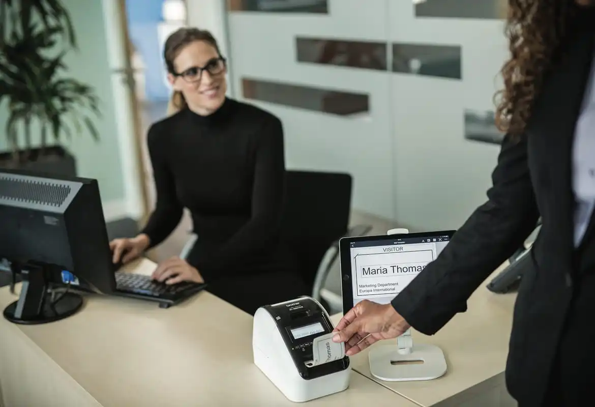 A lady removing a visitor name badge from a Brother QL-810W printer at an office reception desk