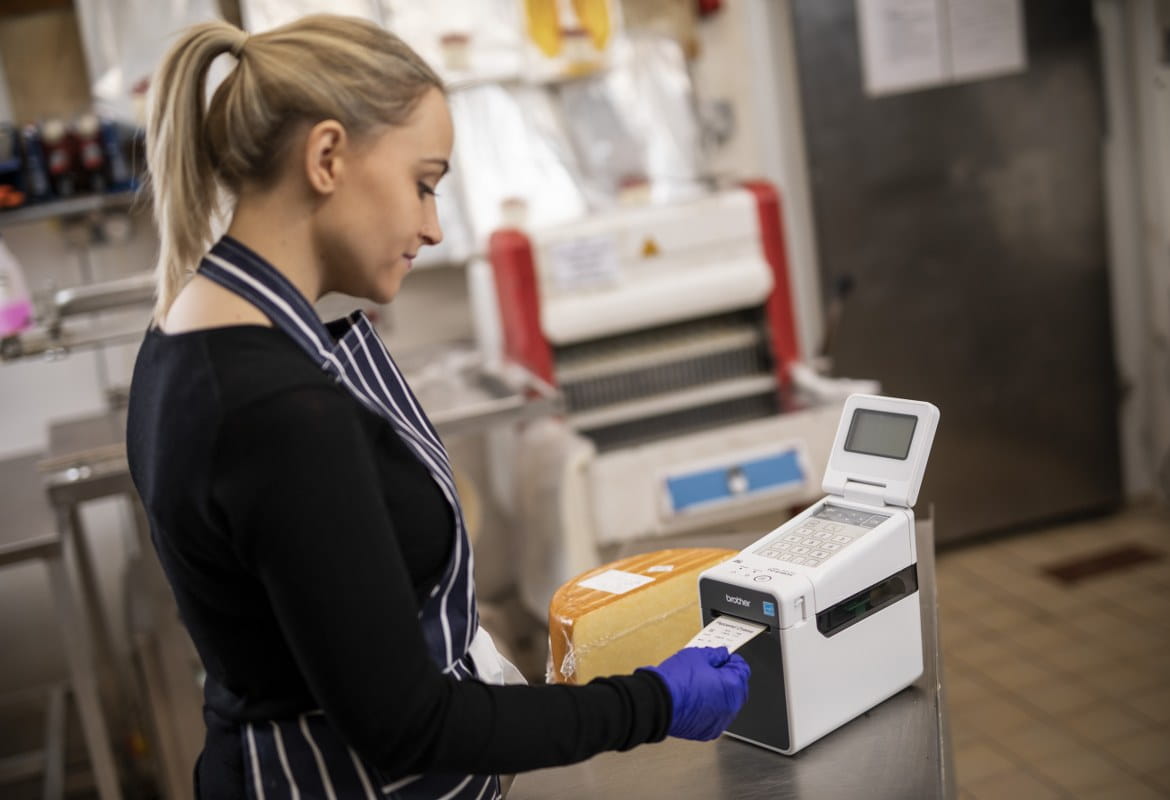 A female member of staff is printing a food label from a Brother labelling machine to add to a wheel of cheese in the back office of a food deli or cafe.