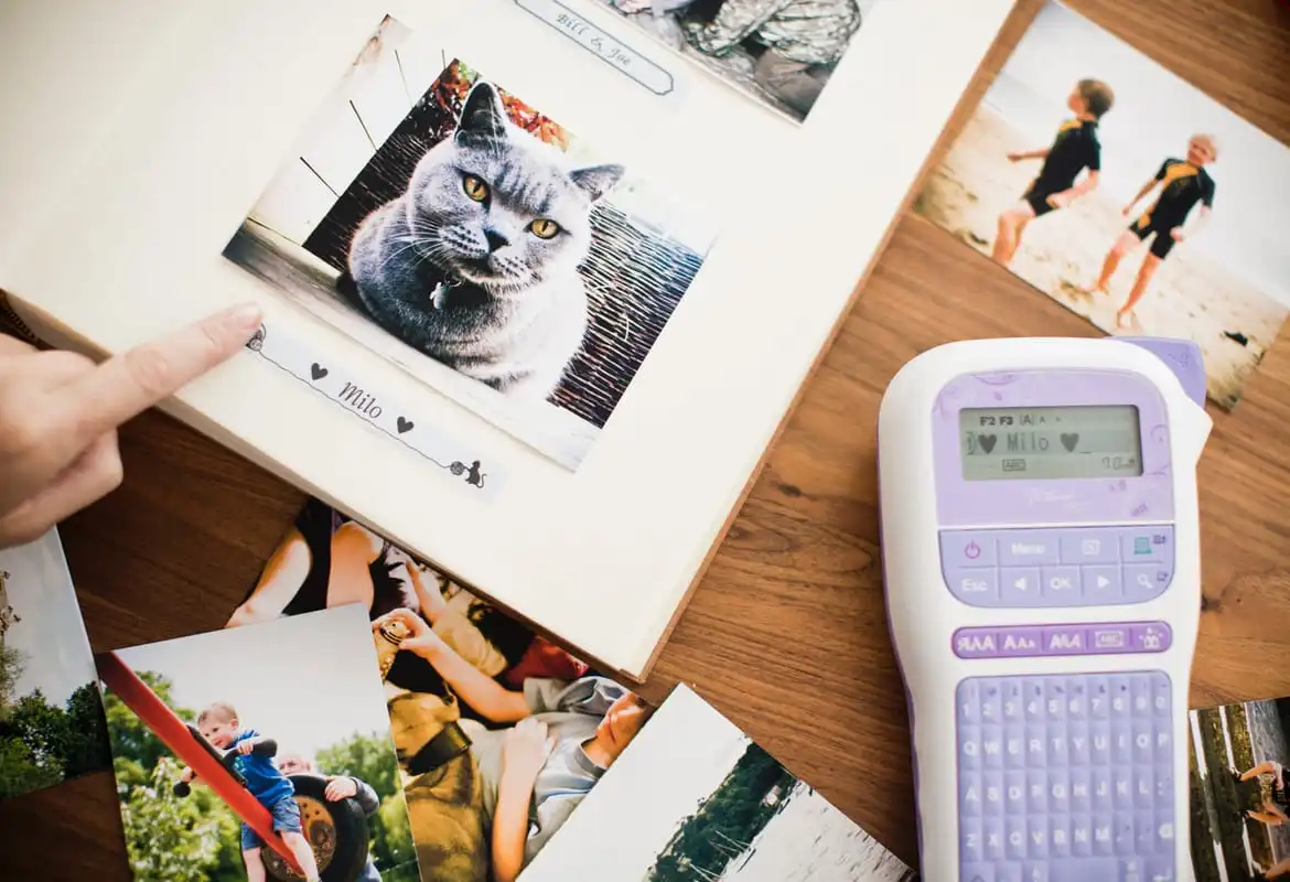 A person applying a sticker to a scrapbook underneath a picture of a cat with a Brother PT-H200 label printer to the side