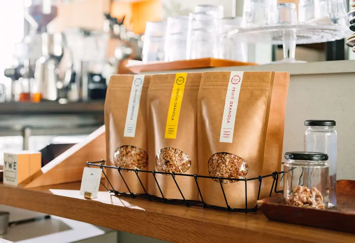 Three brown paper bags containing granola with branded colour stickers on a counter top in a deli