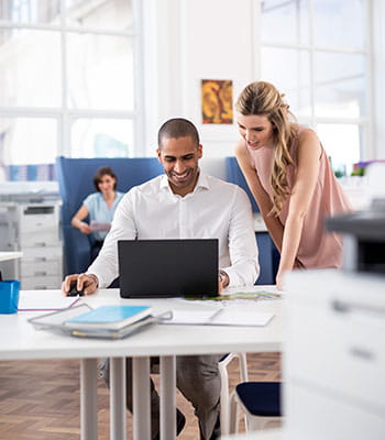 Femme portant des lunettes se tenant debout avec une tasse regardant le pc de son collègue assis à un bureau, imprimantes, homme en costume, ordinateurs, papier