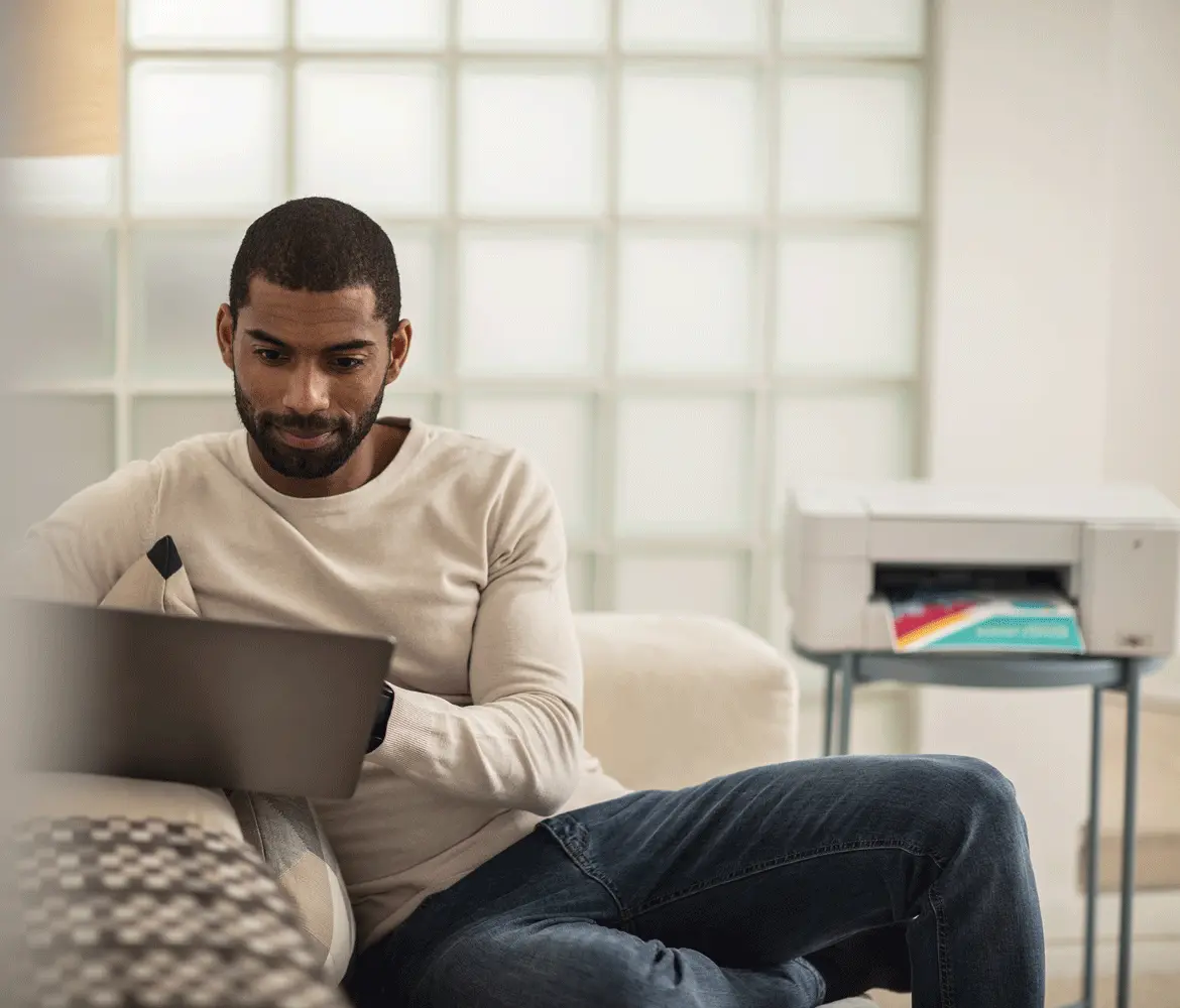 A man sat on a sofa using a laptop with a Brother printer in the background