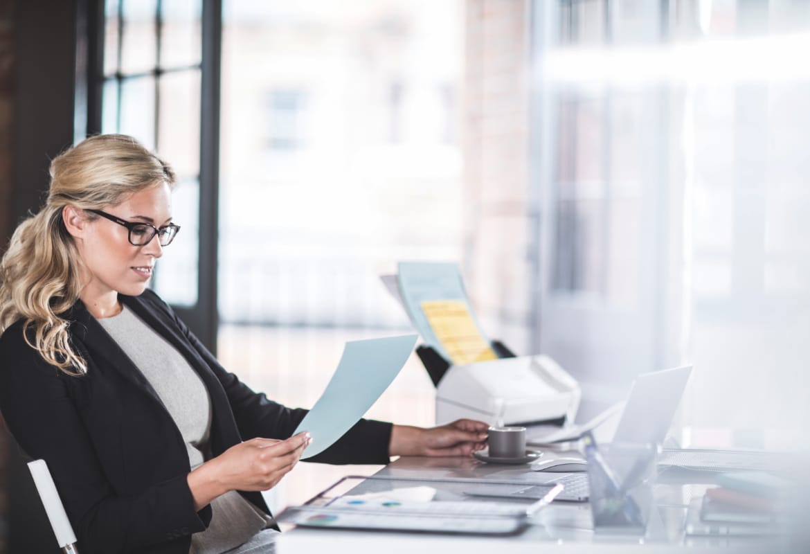 A business woman reading a document while sitting at a desk with a scanner to her side in an office environment