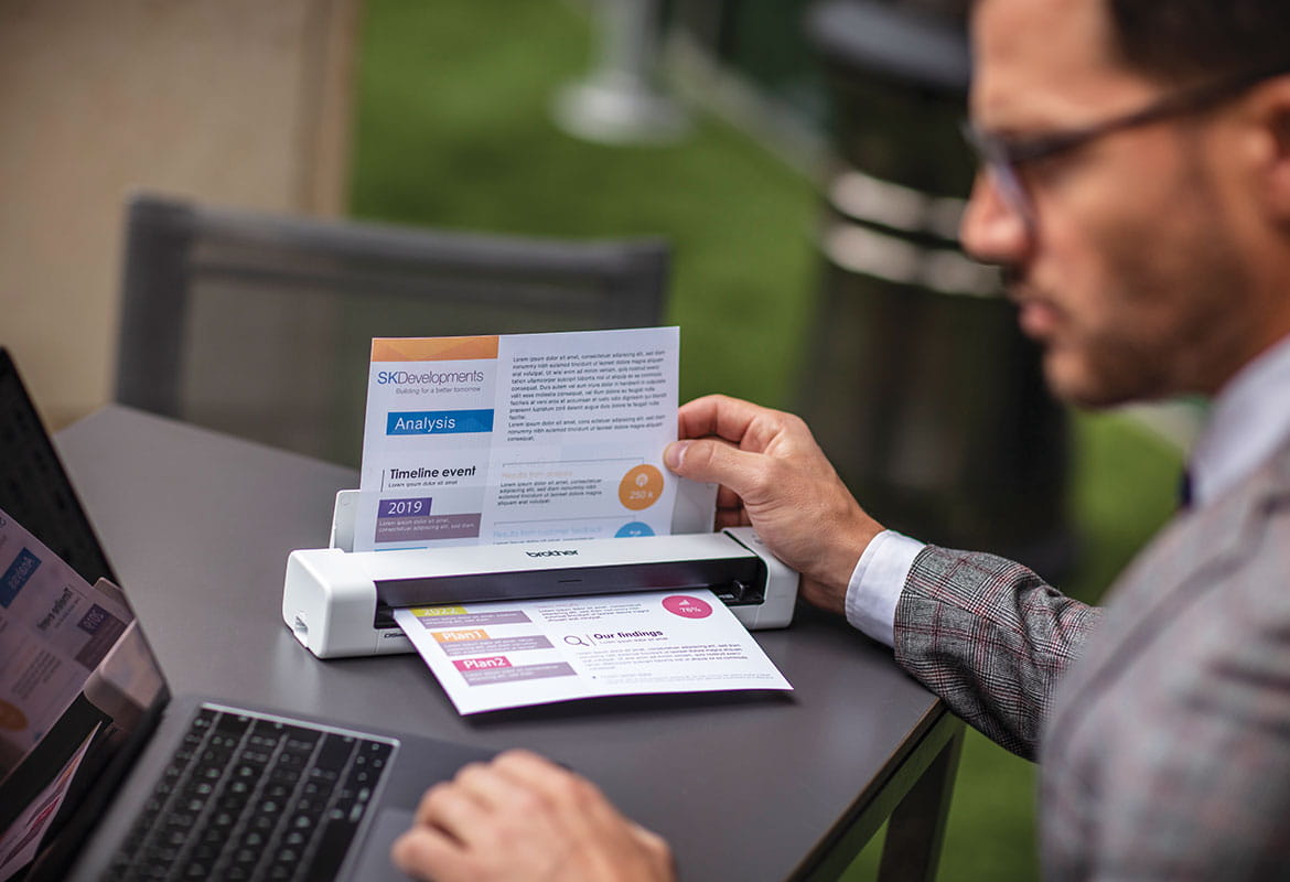 Male sat at grey table wearing glasses, grey suit with laptop scaniing colour document outdoors