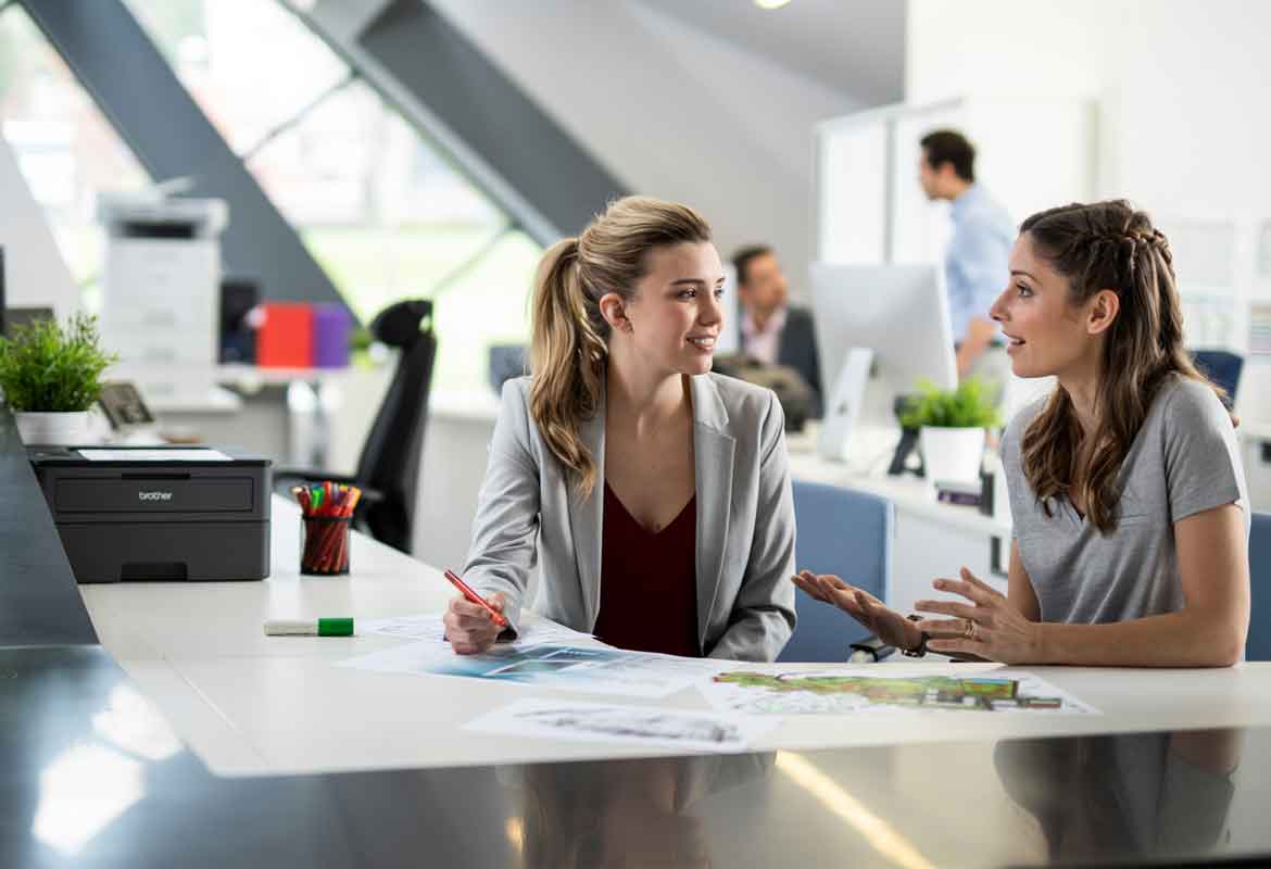 Two woman sat in a busy office at a desk talking, men in the background, printers, pens, pen pot, documents