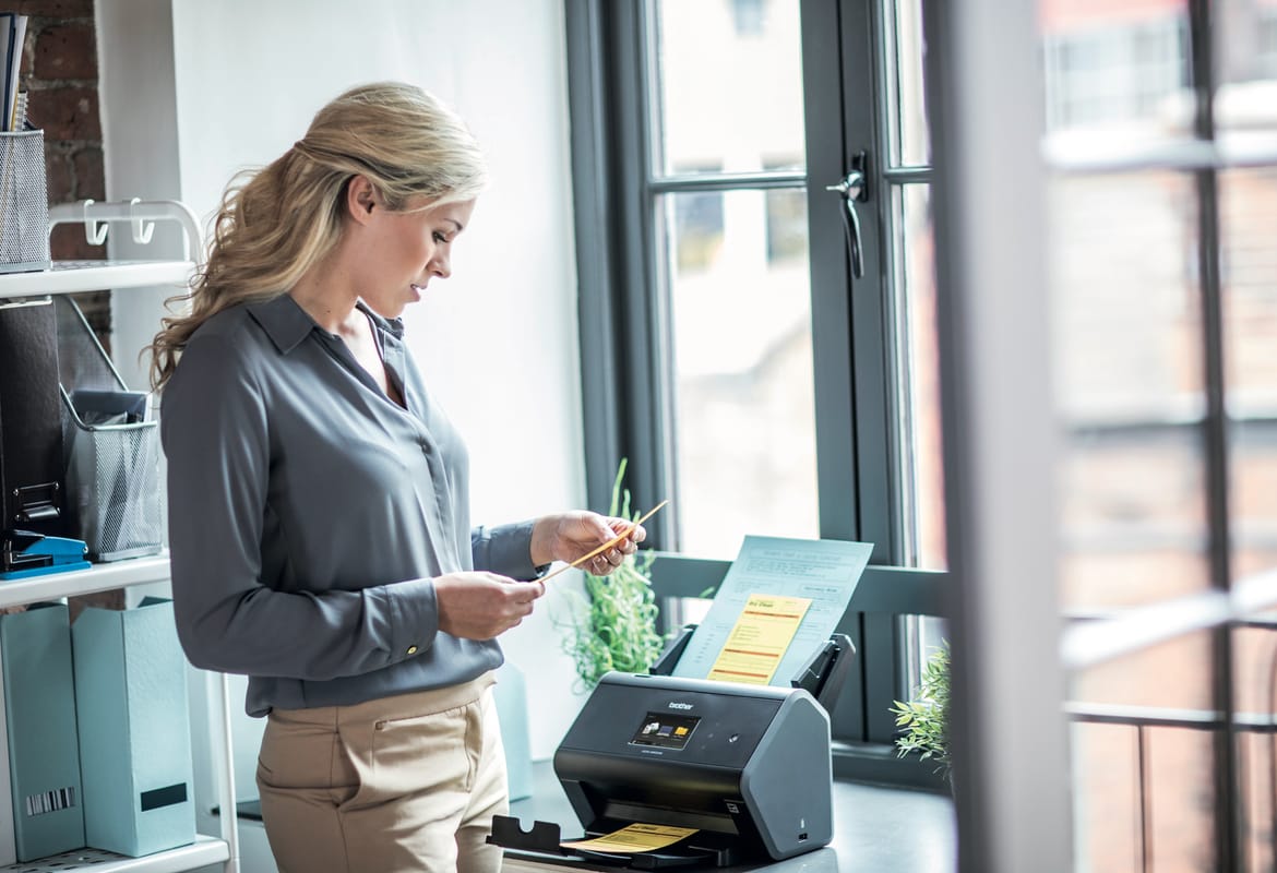 A business woman scanning documents using a Brother ADS-2800W desktop document scanner in a small office environment