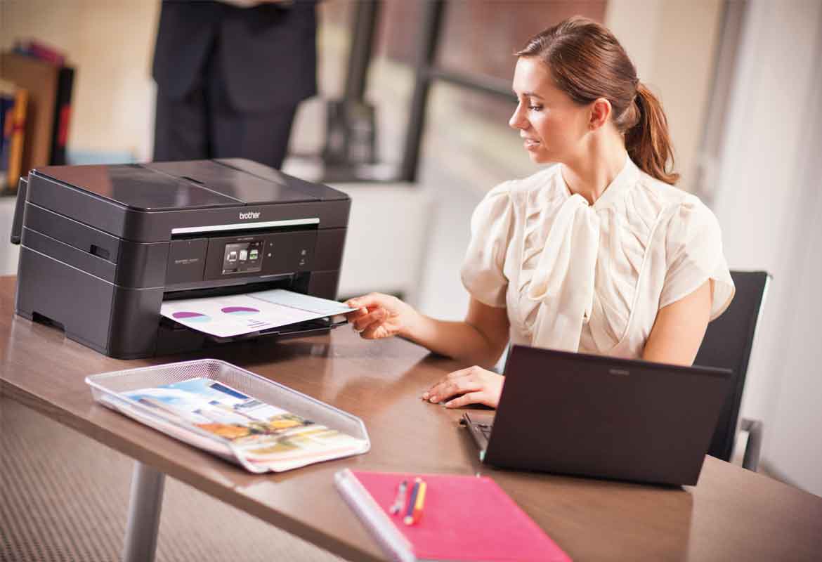 A lady working on a laptop and taking paper off the printer on her desk