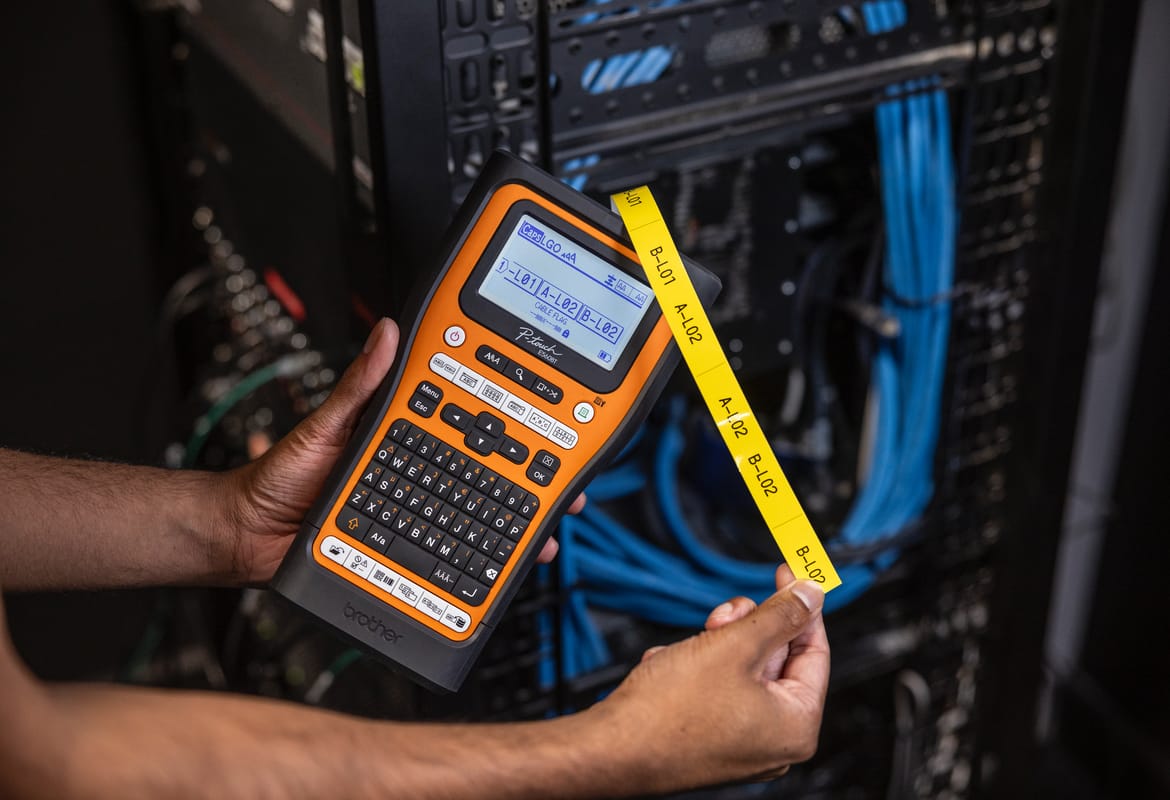 Close-up of an electrician using a Brother P-touch E-series label printer to print an electrical label in front of a network rack unit