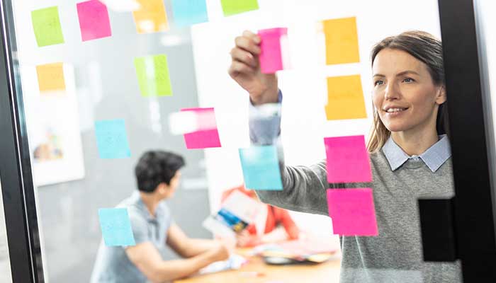 Woman putting colour post-it notes on glass, man and woman in background, colour documents, table