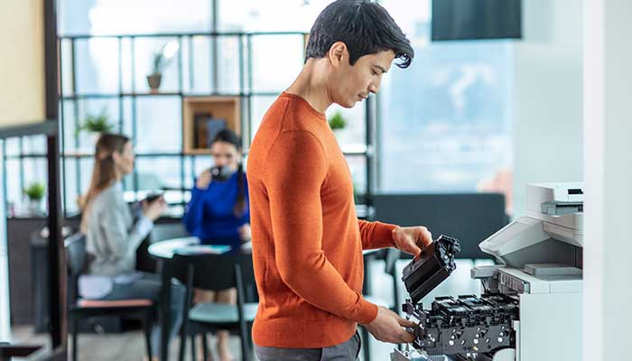 Man wearing orange holding toner near printer, women sat a table