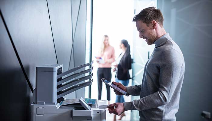 Man holding NFC card near printer, holding tablet, people in background