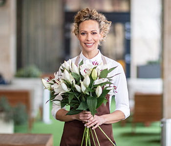 Une fleuriste portant un tablier marron et ayant les cheveux blonds bouclés tient un bouquet de lys blanc.