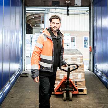 Homme portant un manteau de sécurité orange tirant un transpalette avec des cartons posés dessus dans un camion aux parois bleus