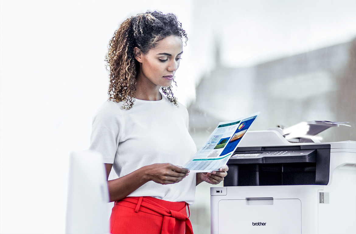 A woman reviewing a printed piece of paper whilst standing next to a Brother printer