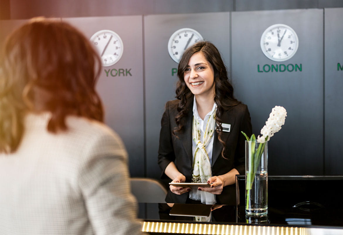 Hotel guest at reception with concierge, clocks, computer, vase