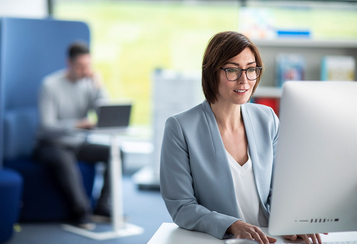 Woman wearing glasses working on computer in office with printer and man using laptop