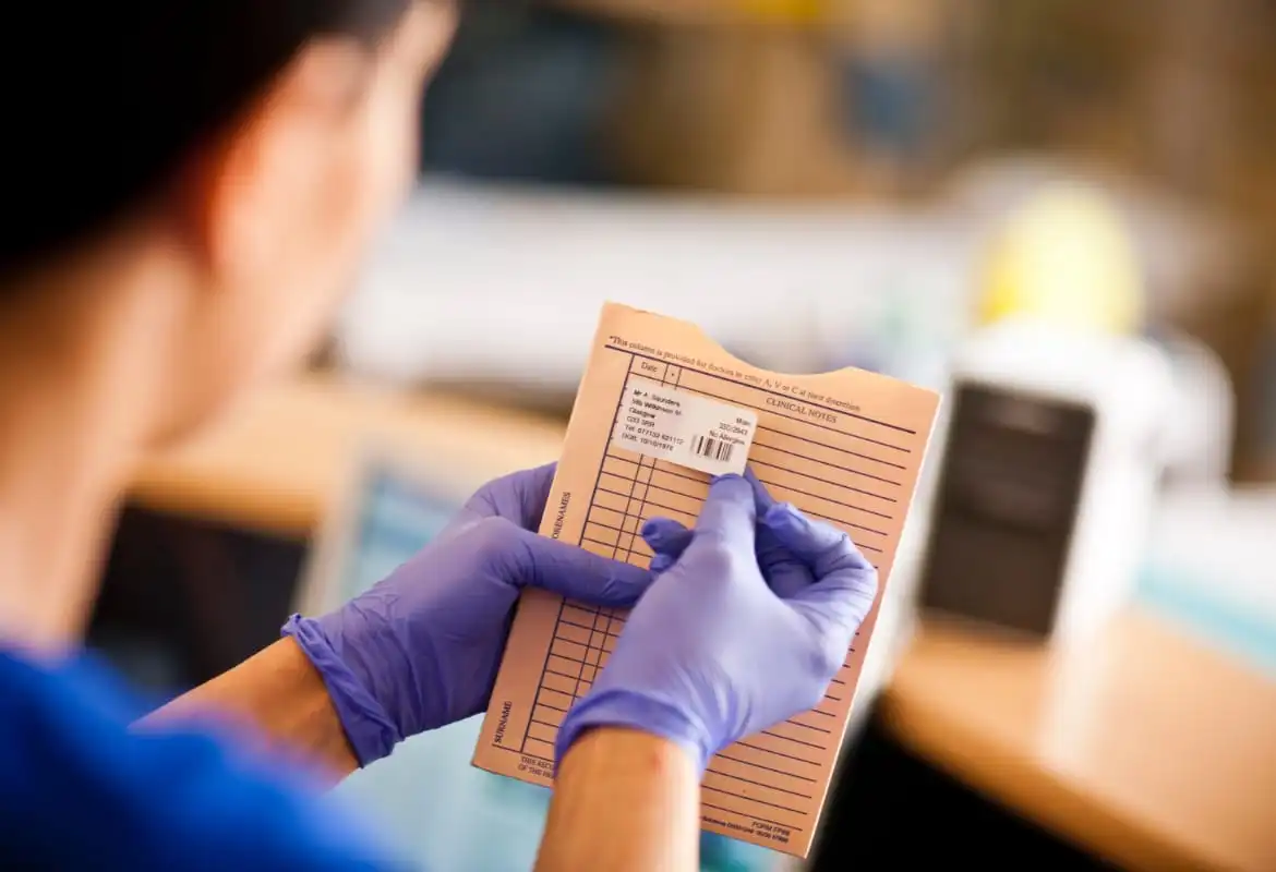 Male doctor with blue gloves holding patient file with patient details on label, female patient lying down on bed with medical equipment