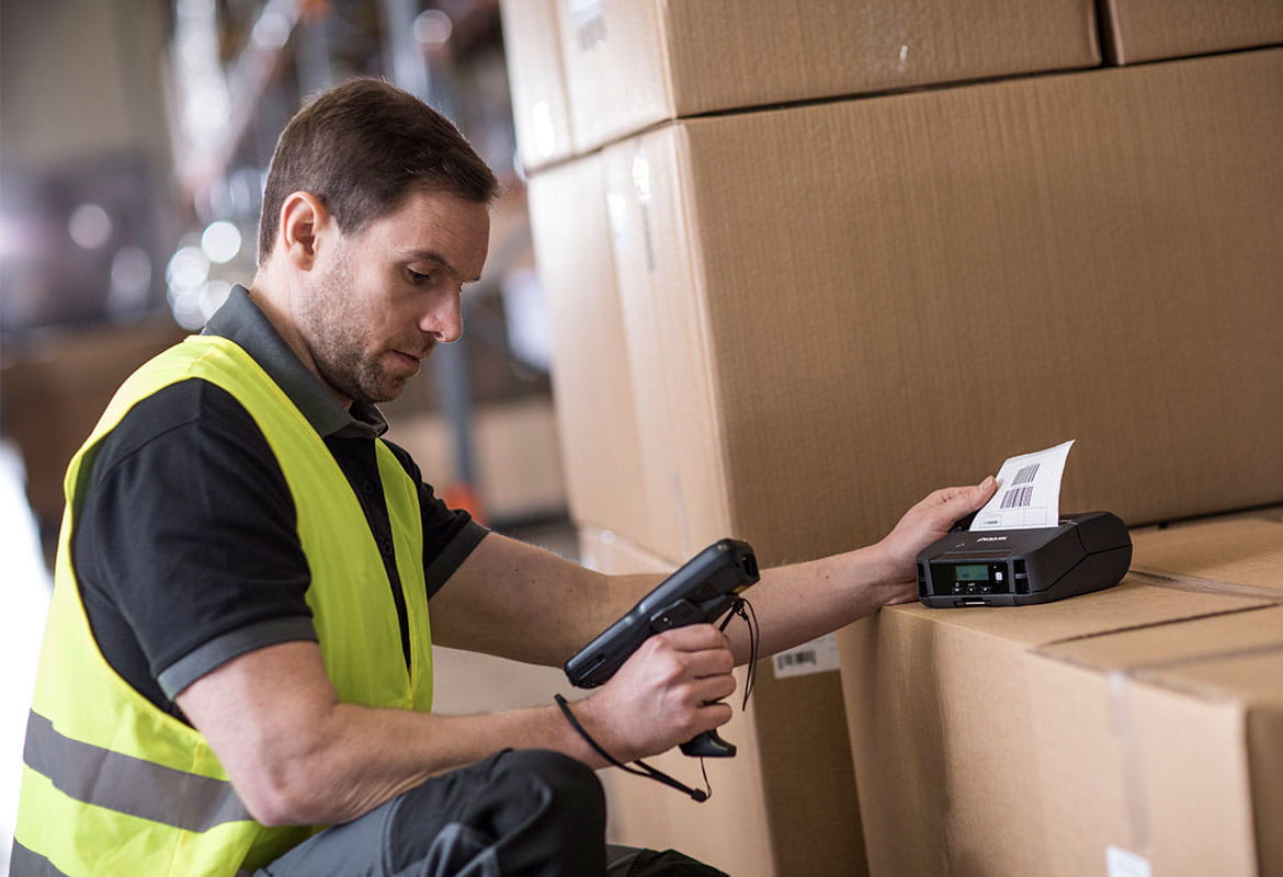 Man wearing hi-vis holding scanner printing label, label printer on top of boxes in warehouse