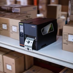 A brother industrial label printer on a shelf surrounded by brown cardboard boxes in a warehouse environment