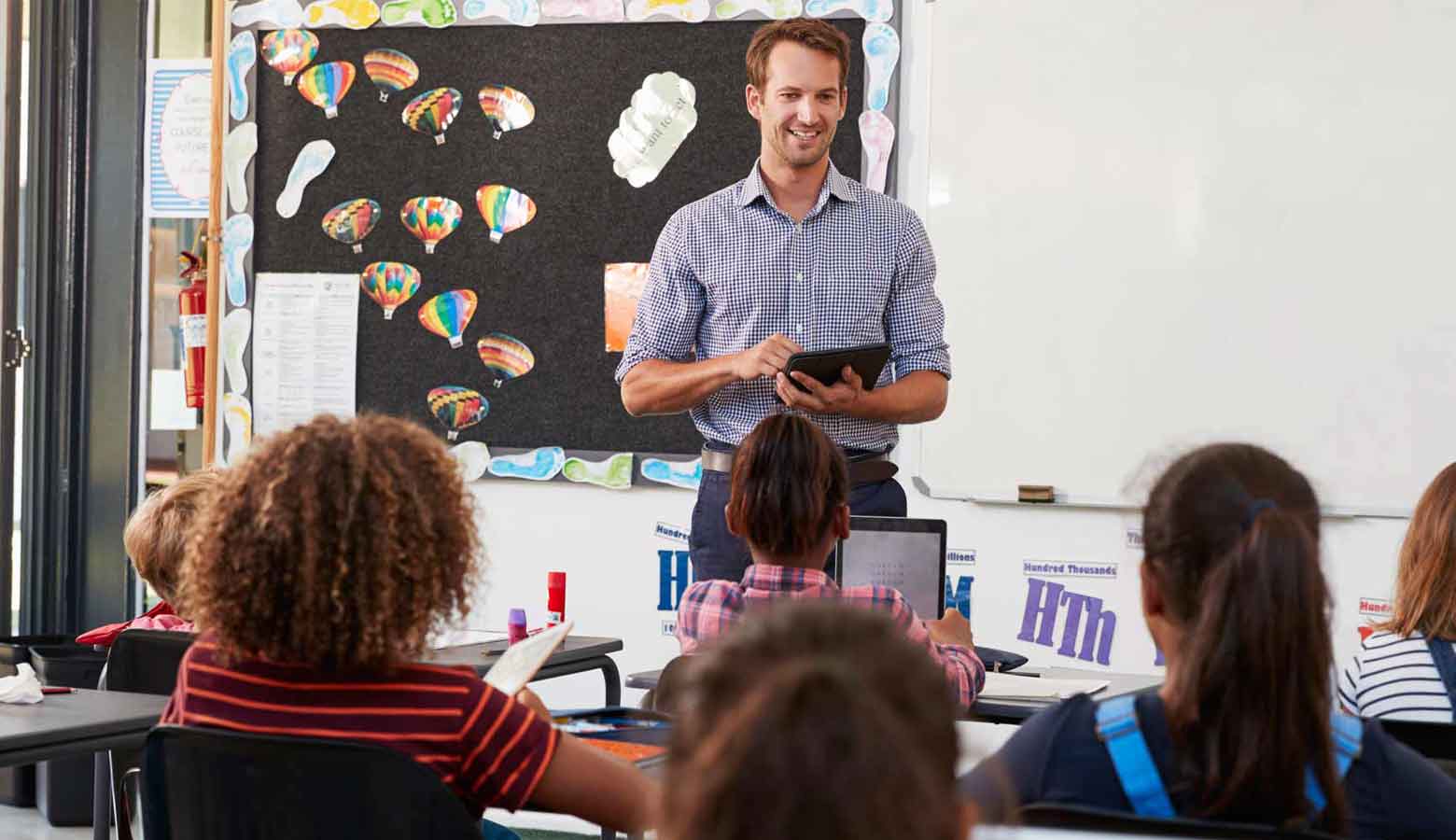 A male teacher with a tablet in his hand speaking to children in the classroom