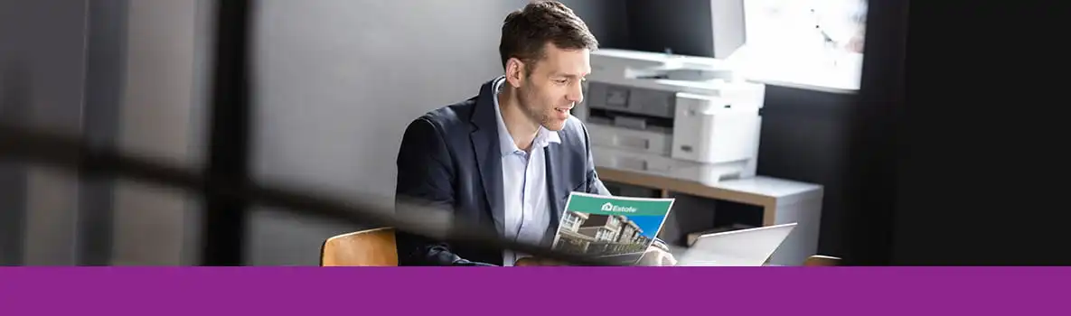 Un homme devant son ordinateur tient dans sa main un document. Derrière lui une imprimante Brother est posée sur un meuble.