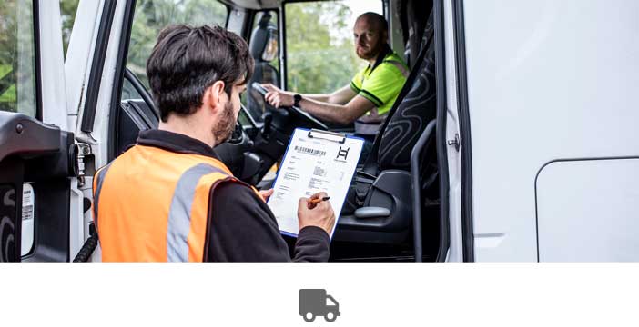 Un homme assis dans un camion, porte ouverte, un homme en veste de sécurité orange se tient debout devant lui avec un presse-papiers dans les mains