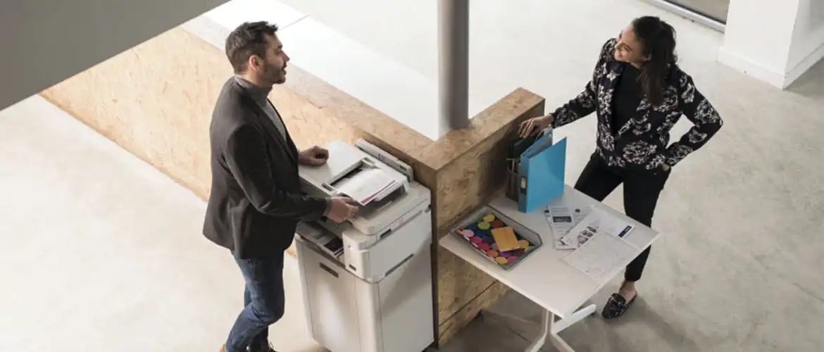 Homme attendant la sortie de son impression sur une imprimante multifonction Brother et parlant avec une femme adossé à un bureau en face en train de discuter ensemble