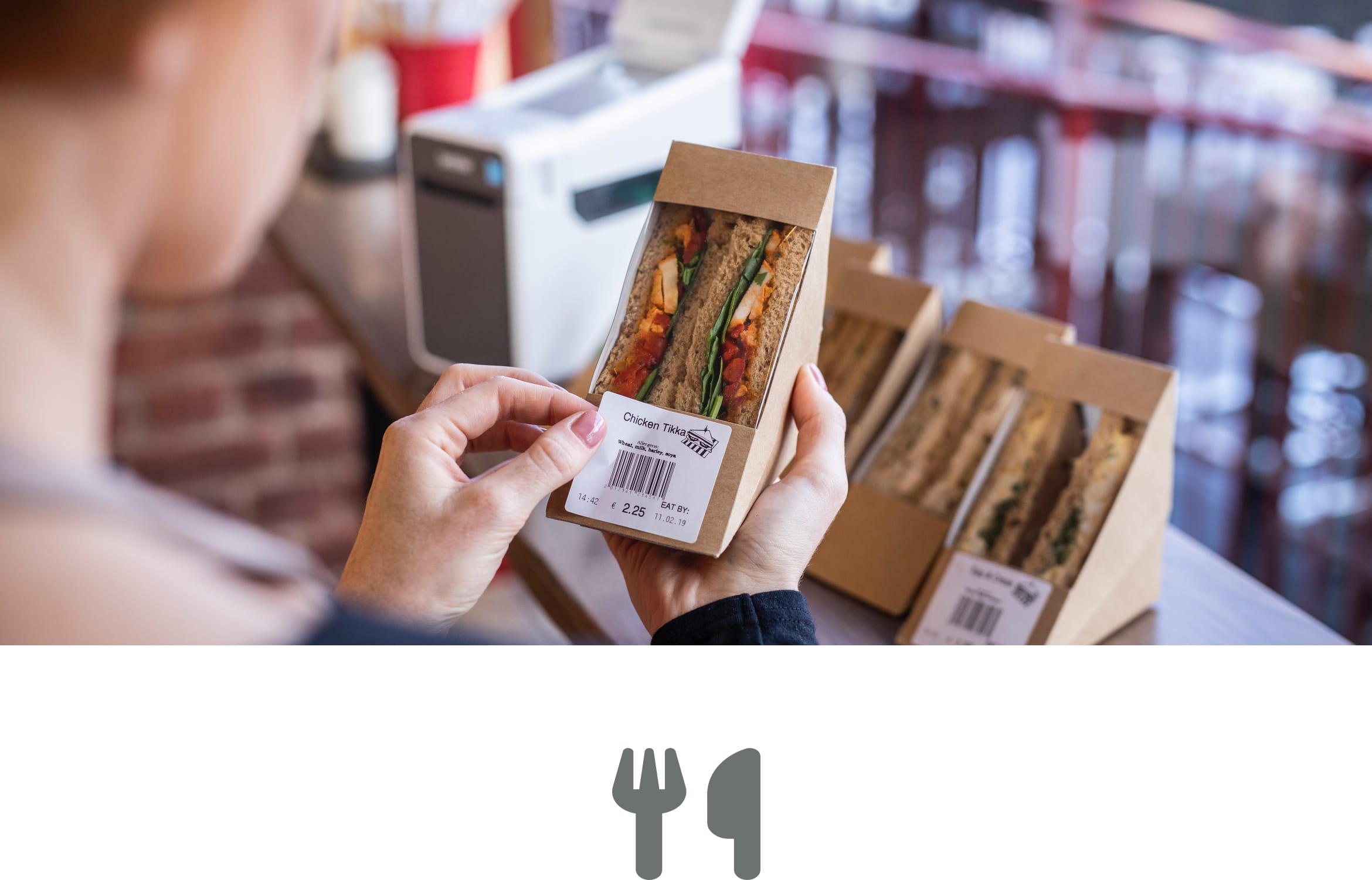 A female sales assistant applying a barcode label to a sandwich box at a counter in a high street deli