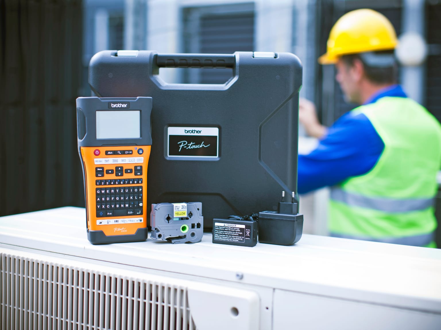 Brother PT-E550 labeller with supplies and accessories on a commercial air conditioning unit outside a building with an electrician wearing a hard hat and high visibility vest in the background
