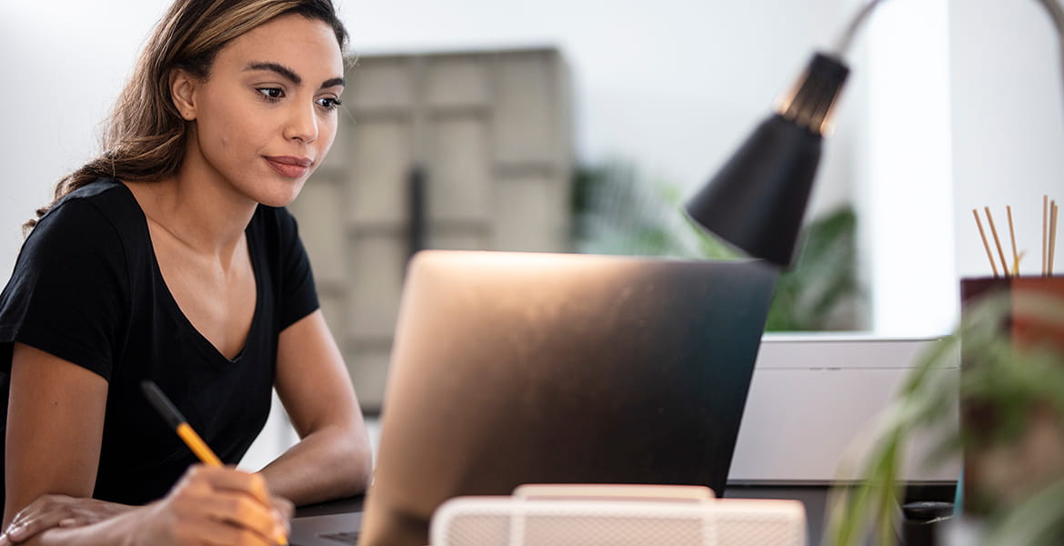 Femme tenant un stylo et regardant un ordinateur portable avec une lampe posée sur le bureau