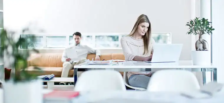 Femme assise à un bureau avec un ordinateur portable, homme assis au fond sur un canapé orange, plantes, cahiers, chaises blanches, fenêtre